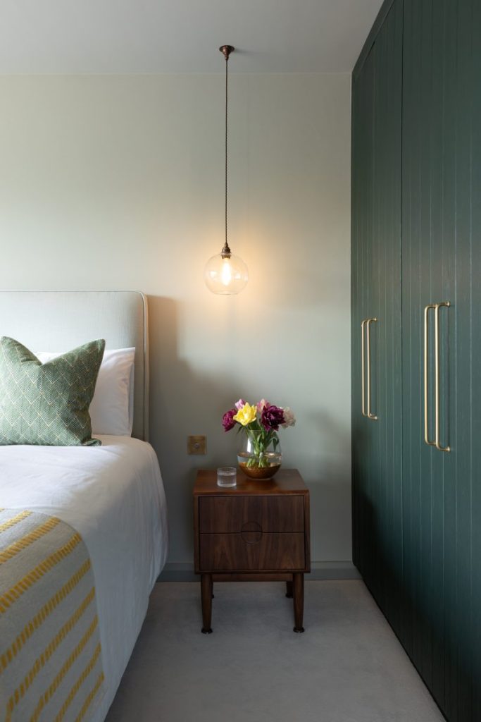 Minimalist bedroom corner with wooden bedside table, pendant light, and upholstered bed against a muted green wall.