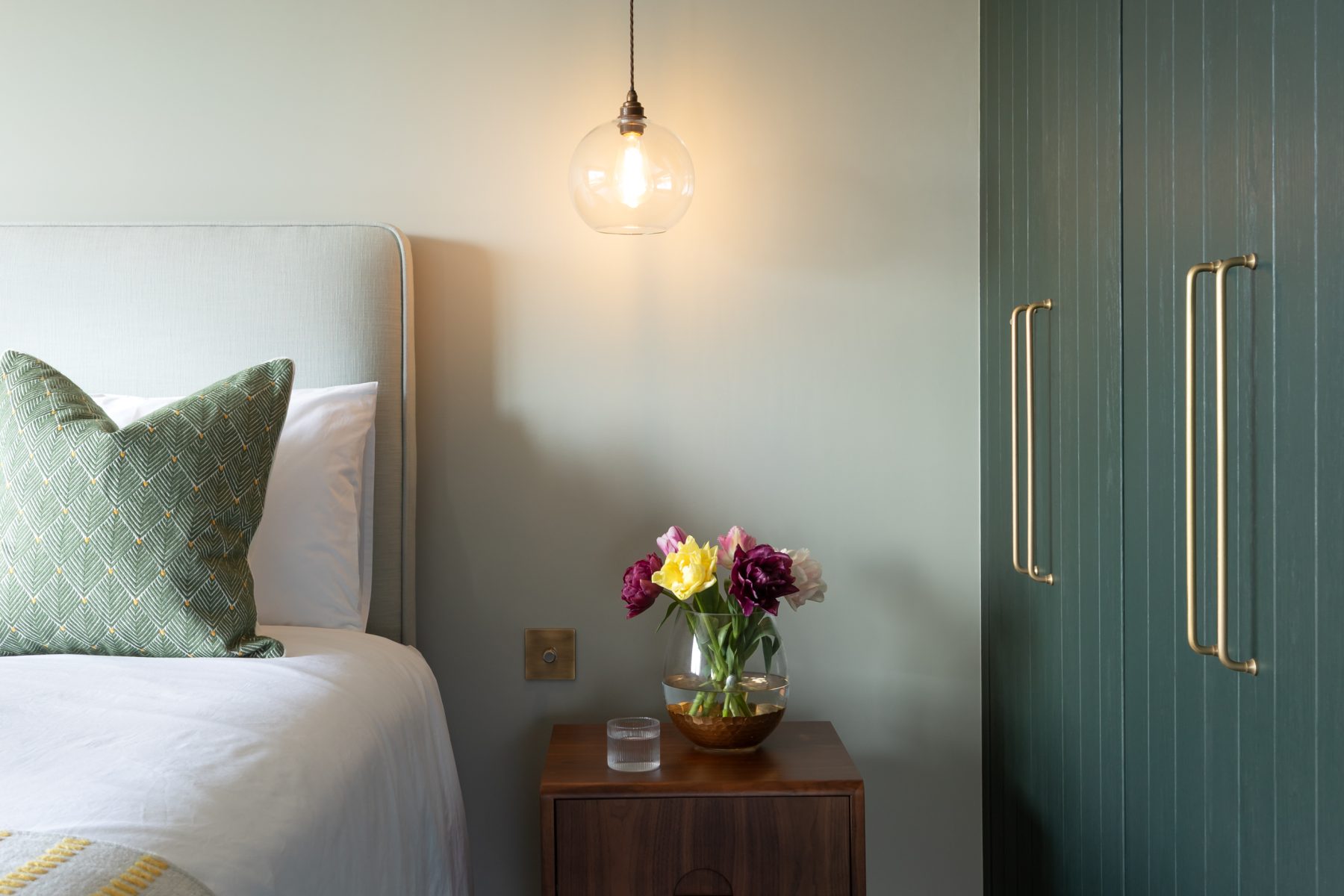 Minimalist bedroom corner with wooden bedside table, pendant light, and upholstered bed against a muted green wall.