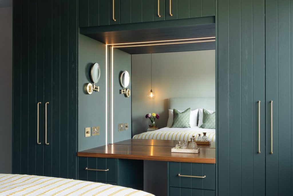 Dark green fitted wardrobes with integrated dressing table, illuminated mirror, and brass handles in a contemporary bedroom.