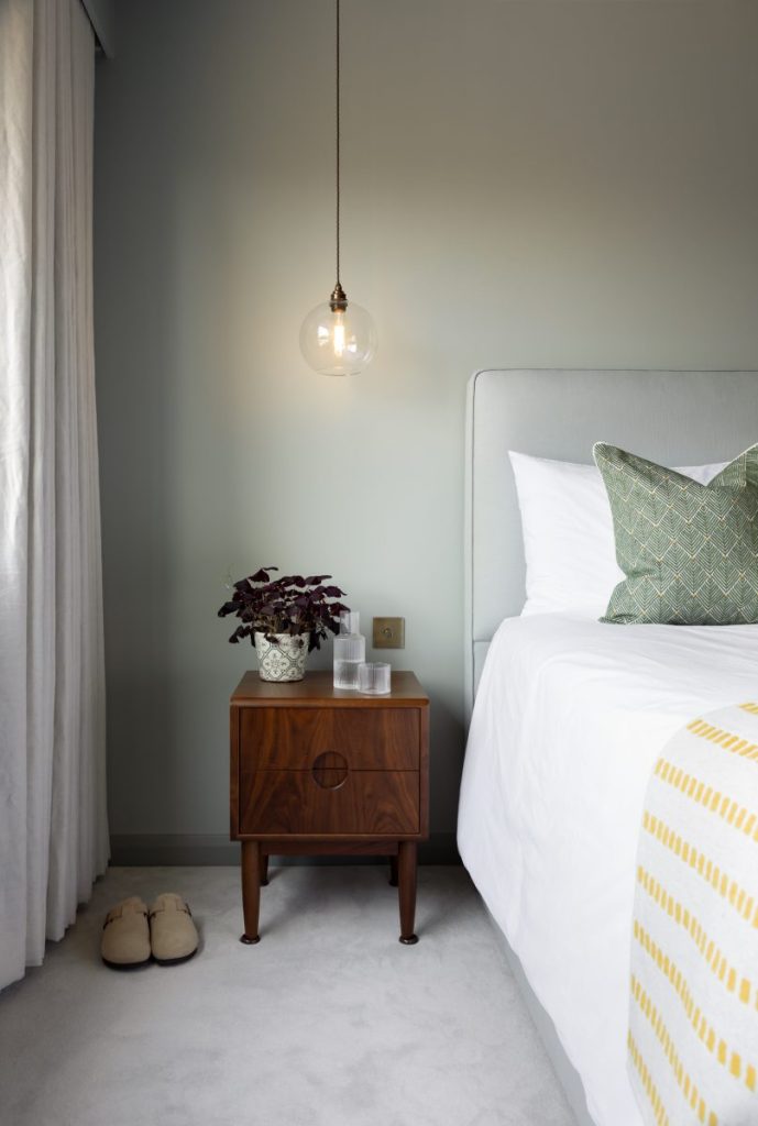 Minimalist bedroom corner with wooden bedside table, pendant light, and upholstered bed against a muted green wall.