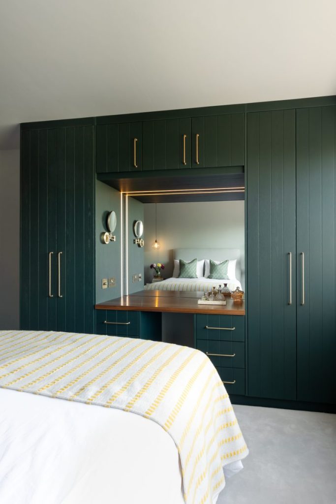 Dark green fitted wardrobes with integrated dressing table, illuminated mirror, and brass handles in a contemporary bedroom.