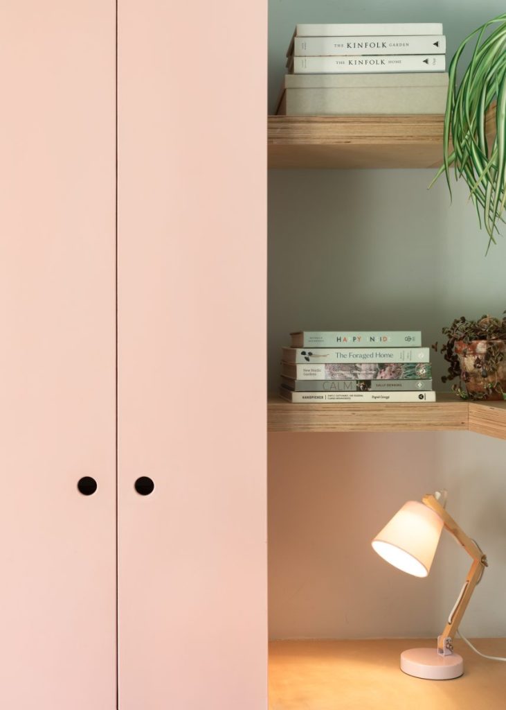 Soft-toned built-in shelving with books, decorative objects, and modern desk lamp beside blush cabinetry.