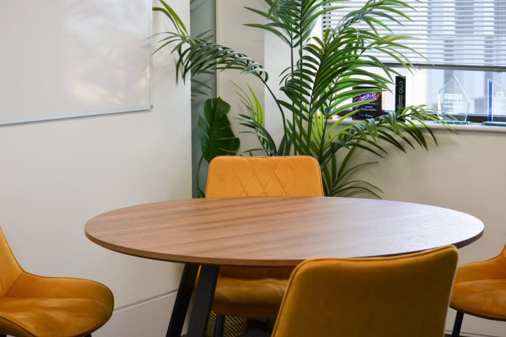 Round timber meeting table with mustard upholstered chairs and indoor plants in contemporary workspace.