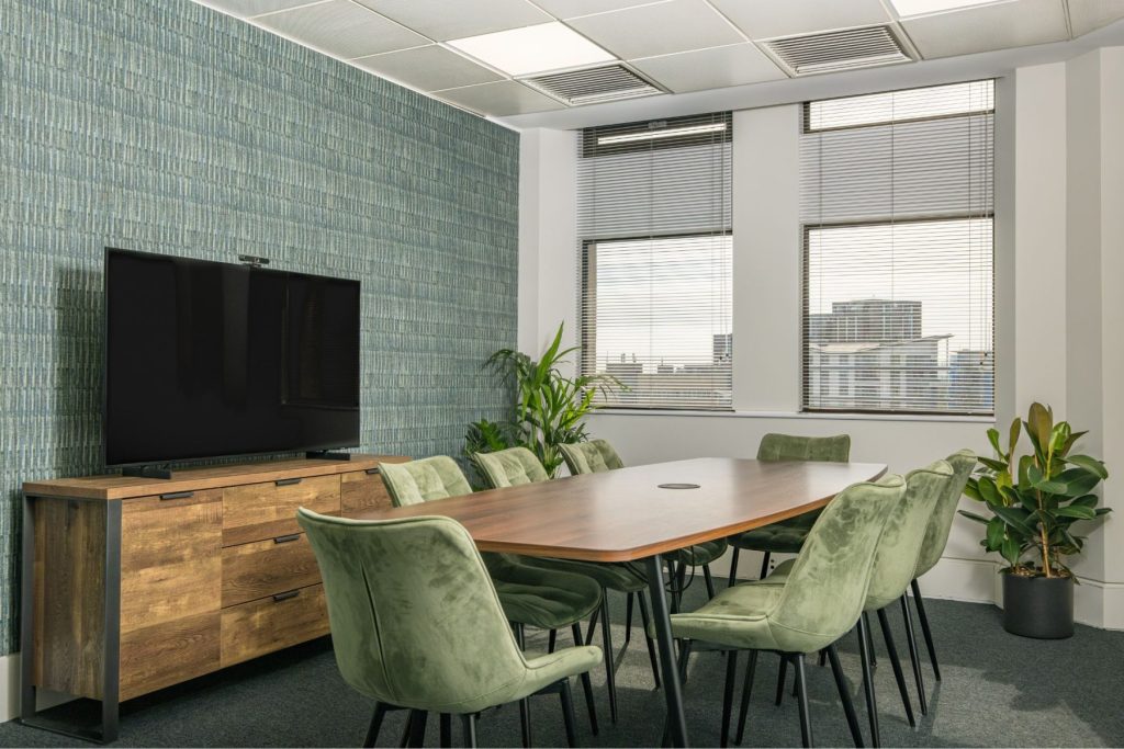 Contemporary meeting room with woodconference table, green upholstered chairs, and large display screen.