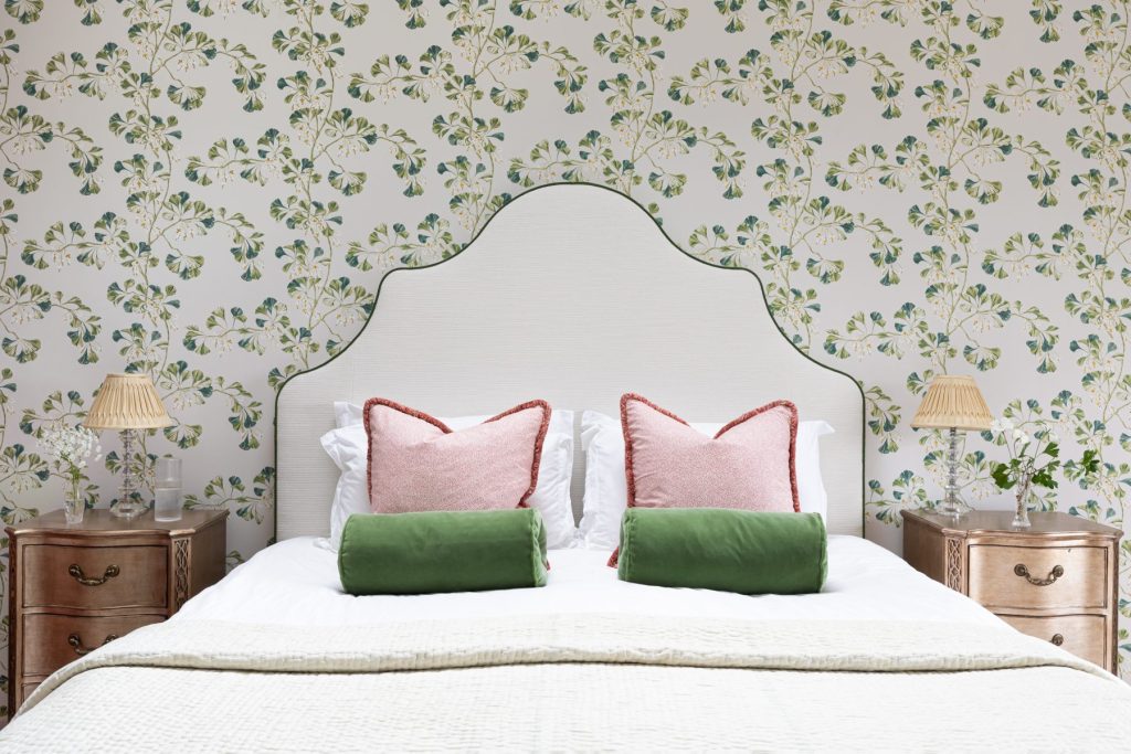 Bedroom with upholstered headboard, floral wallpaper, wooden bedside tables and layered cushions in soft pink and green tones.