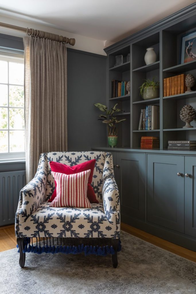 Built-in blue cabinetry with shelving, neutral curtains and an upholstered chair creating a calm reading corner in a residential interior.
