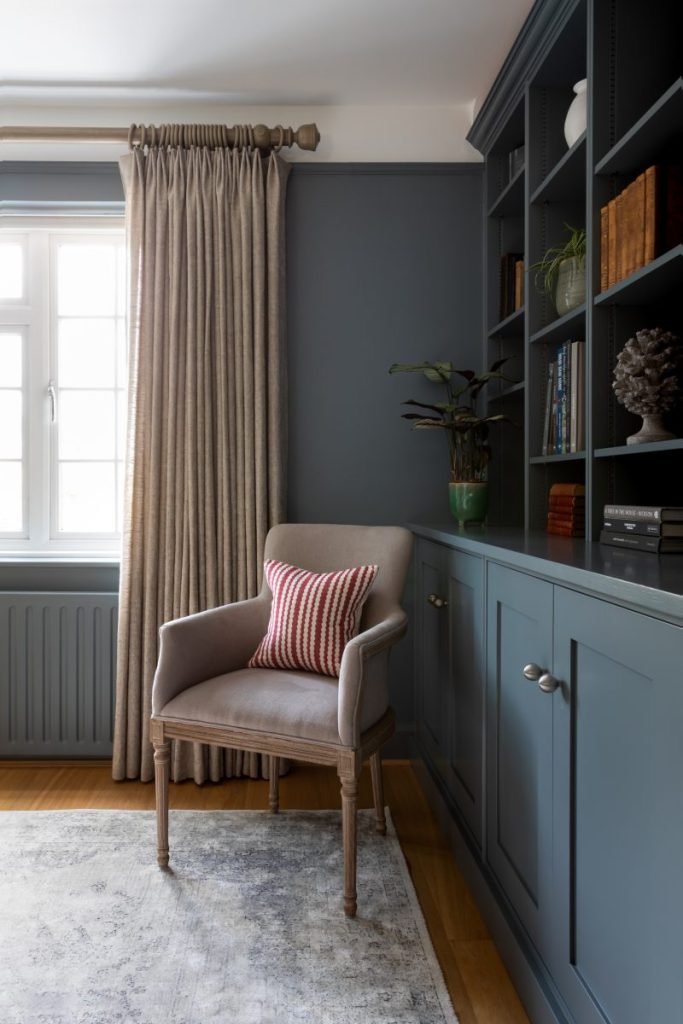 Built-in blue cabinetry with shelving, neutral curtains and an upholstered chair creating a calm reading corner in a residential interior.