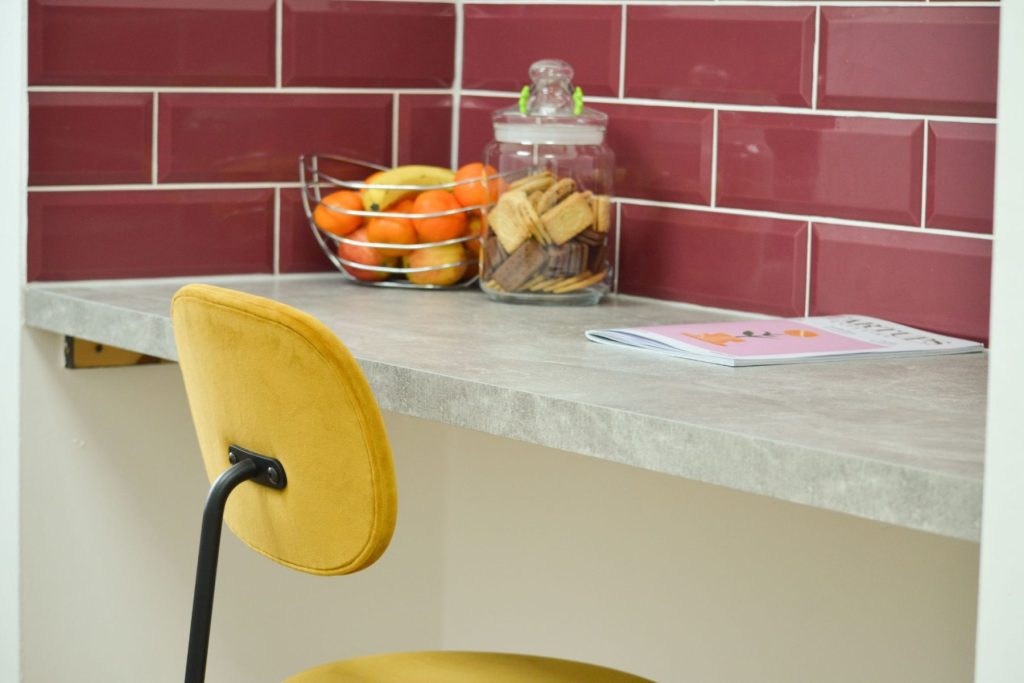Modern office breakout area with red tiled backsplash, concrete-effect worktop, and mustard upholstered chair.”