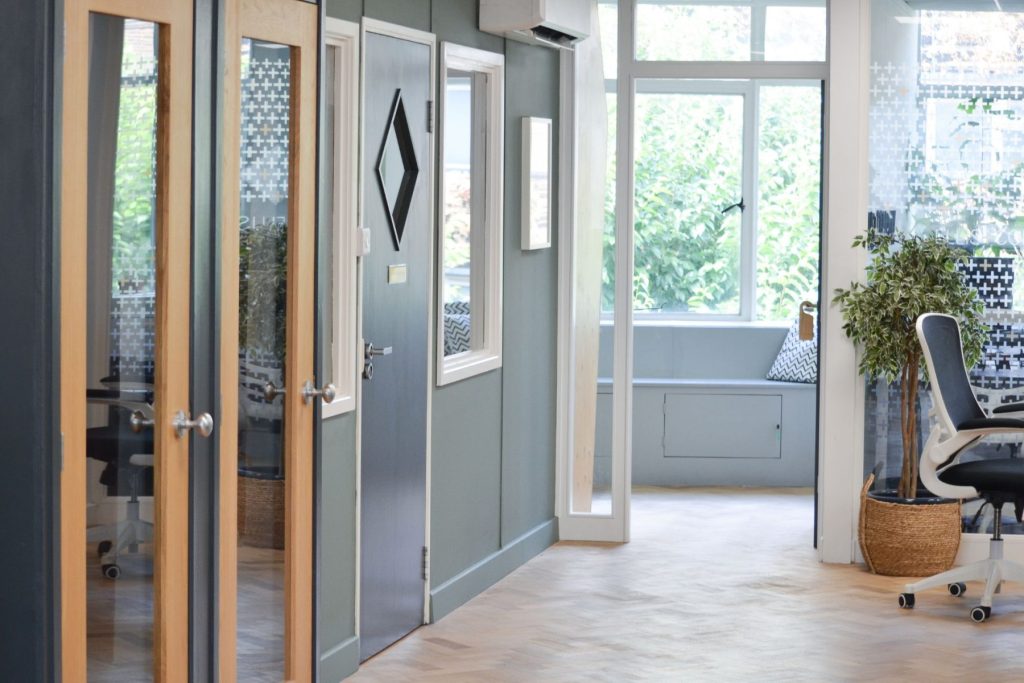 Modern office interior with glass-fronted rooms, wooden doors, herringbone flooring, and indoor plant.