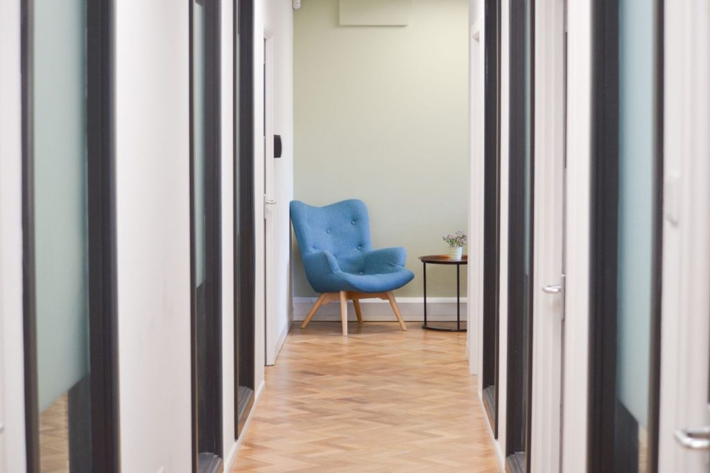 Modern office corridor with glass partitions, herringbone flooring, and upholstered lounge chair.”