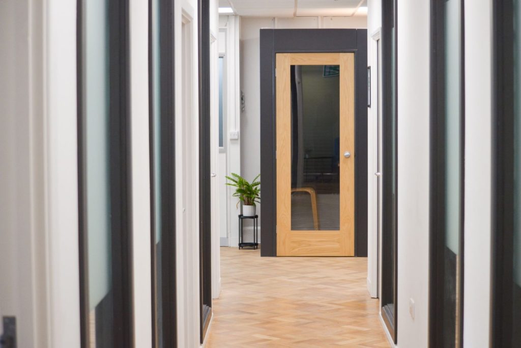 Modern office corridor with glass partitions, wooden door, herringbone flooring, and indoor plant.