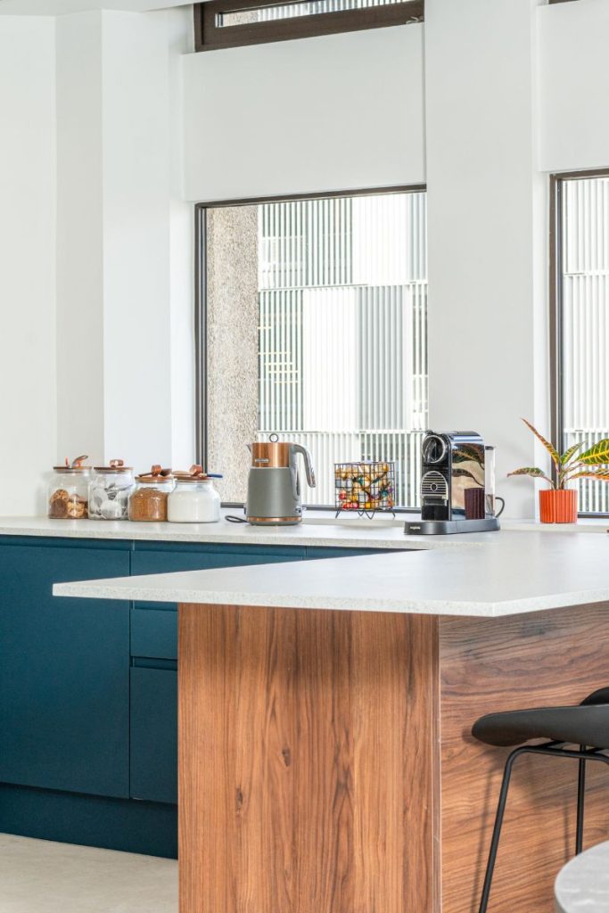 Modern office kitchenette with wooden island, blue cabinetry, coffee machine, and large windows.