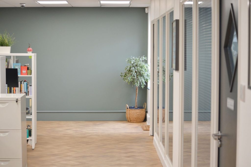 Modern office interior with glass partitions, herringbone flooring, muted wall tones, and indoor plant.
